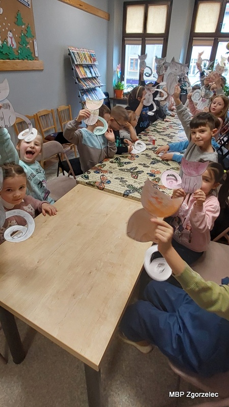 Dzieci siedzą przy długim stole w&nbsp;bibliotece i&nbsp;trzymają w&nbsp;rękach papierowe kotki wykonane podczas zajęć plastycznych. Stół przykryty jest obrusem z&nbsp;motylami, a&nbsp;w tle widoczne są regały z&nbsp;książkami oraz stojaki z&nbsp;publikacjami. Dzieci podnoszą swoje prace w&nbsp;górę, prezentując różnokolorowe rysunki kotów o&nbsp;spiralnych ogonach. Zdjęcie wykonano 24 lutego 2026 r. w&nbsp;Oddziale dla Dzieci i&nbsp;Młodzieży.