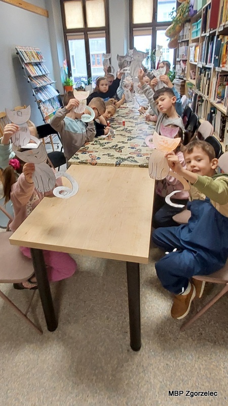 Dzieci siedzą przy długim stole w&nbsp;bibliotece i&nbsp;trzymają w&nbsp;rękach papierowe kotki wykonane podczas zajęć plastycznych. Stół przykryty jest obrusem z&nbsp;motylami, a&nbsp;w tle widoczne są regały z&nbsp;książkami oraz stojaki z&nbsp;publikacjami. Dzieci podnoszą swoje prace w&nbsp;górę, prezentując różnokolorowe rysunki kotów o&nbsp;spiralnych ogonach. Zdjęcie wykonano 24 lutego 2026 r. w&nbsp;Oddziale dla Dzieci i&nbsp;Młodzieży.