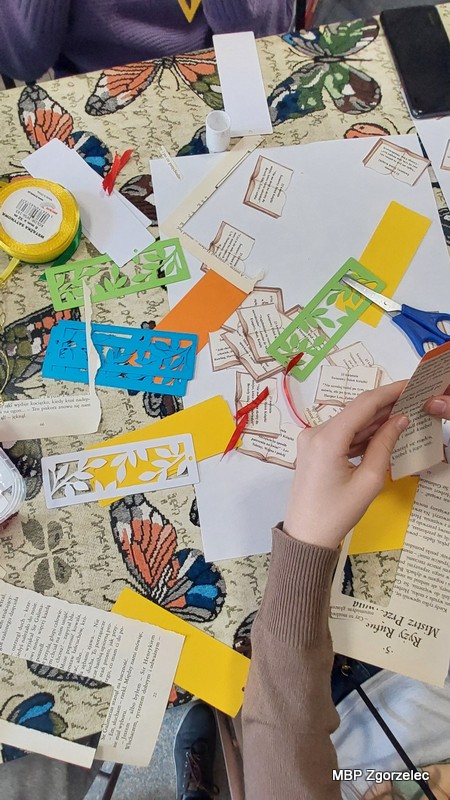 Zbliżenie na stół warsztatowy w&nbsp;bibliotece, na którym znajdują się kolorowe kartony, wycięte wzory, nożyczki, klej i&nbsp;fragmenty stron książek. Widoczne są ręce uczestników wykonujących zakładki do książek w&nbsp;ramach spotkania Młodzieżowego Klubu „Książkowi Wędrowcy”, zorganizowanego z&nbsp;okazji Światowego Dnia Książki i&nbsp;Praw Autorskich.