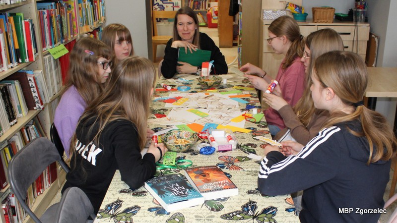 Grupa młodych uczestników siedzi przy stole w&nbsp;Oddziale dla Dzieci i&nbsp;Młodzieży. Na blacie leżą kolorowe kartki, nożyczki, klej oraz gotowe zakładki do książek. W&nbsp;tle widoczne są regały z&nbsp;książkami. Zdjęcie dokumentuje warsztaty towarzyszące spotkaniu klubu „Książkowi Wędrowcy”, zorganizowane z&nbsp;okazji Światowego Dnia Książki.