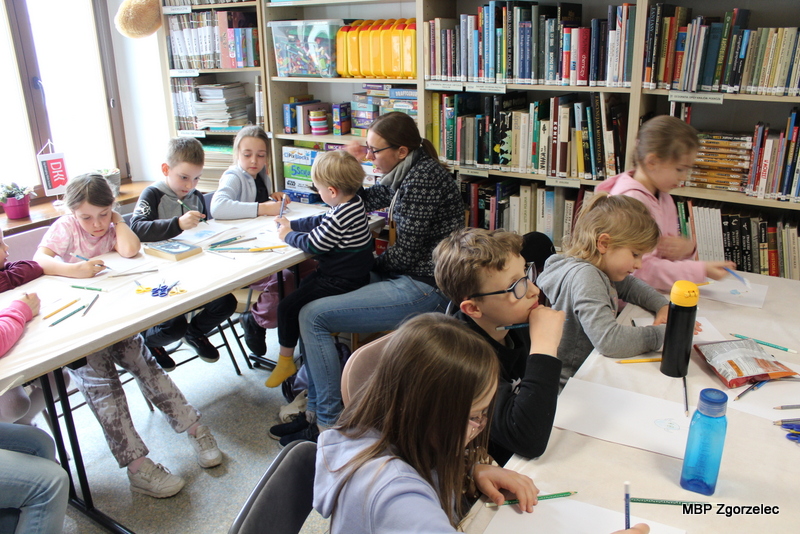 W bibliotecznej sali kilka dzieci i&nbsp;opiekunów siedzi przy długim stole ustawionym między regałami z&nbsp;książkami. Uczestnicy wykonują prace plastyczne związane z&nbsp;tematyką kosmicznej podróży Scottie Go, korzystając z&nbsp;papieru i&nbsp;kredek.
