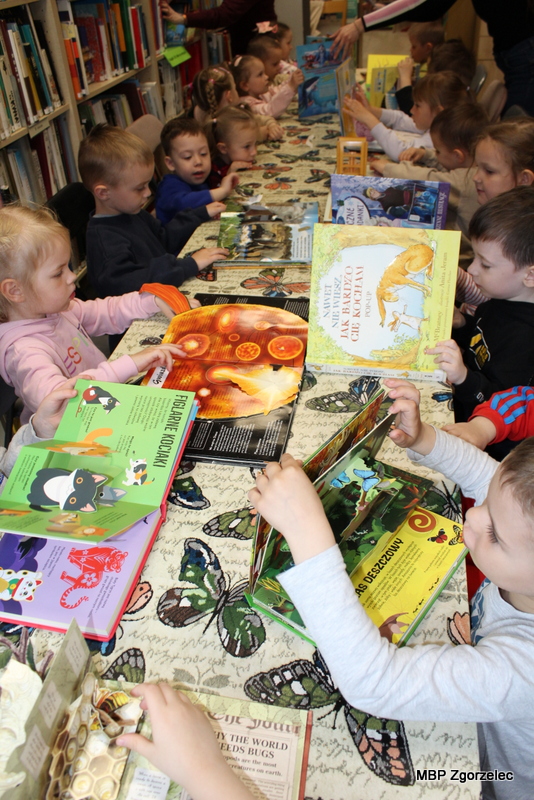Grupa dzieci siedzi przy długim stole w&nbsp;bibliotece. Na stole leżą kolorowe książki obrazkowe, które dzieci oglądają i&nbsp;kartkują. W&nbsp;tle widoczne są regały z&nbsp;książkami, a&nbsp;cała scena przedstawia zajęcia czytelnicze dla najmłodszych.