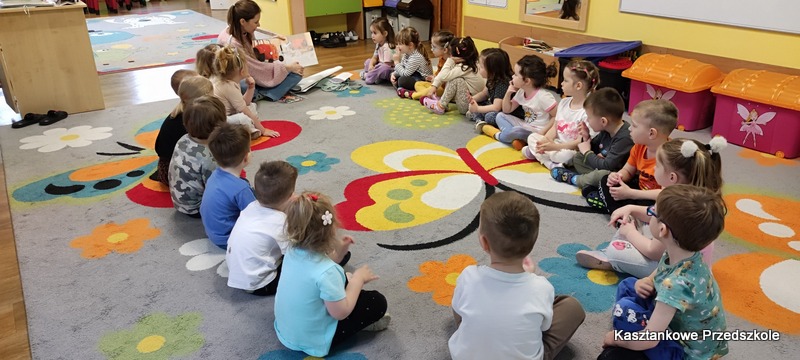 Dzieci siedzą w&nbsp;kręgu na dywanie w&nbsp;sali przedszkolnej i&nbsp;słuchają czytanej książki „Traktorek jest dzielny” podczas zajęć o&nbsp;życiu na wsi.