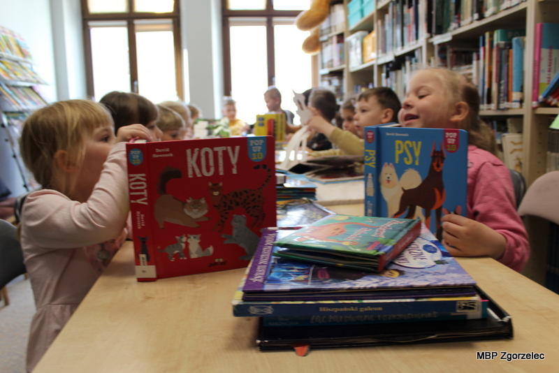 Dzieci siedzące przy stole w&nbsp;bibliotece oglądają książki obrazkowe o&nbsp;zwierzętach. Na pierwszym planie widoczne okładki książek tematycznych, w&nbsp;tle półki z&nbsp;książkami.