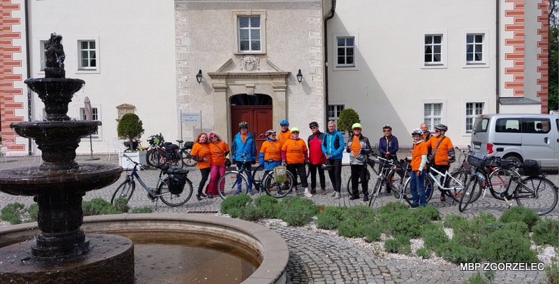 Uczestnicy rajdu rowerowego w&nbsp;pomarańczowych koszulkach stoją przy kamiennej fontannie na dziedzińcu zabytkowego obiektu; w&nbsp;tle widoczna zieleń i&nbsp;zabudowania.