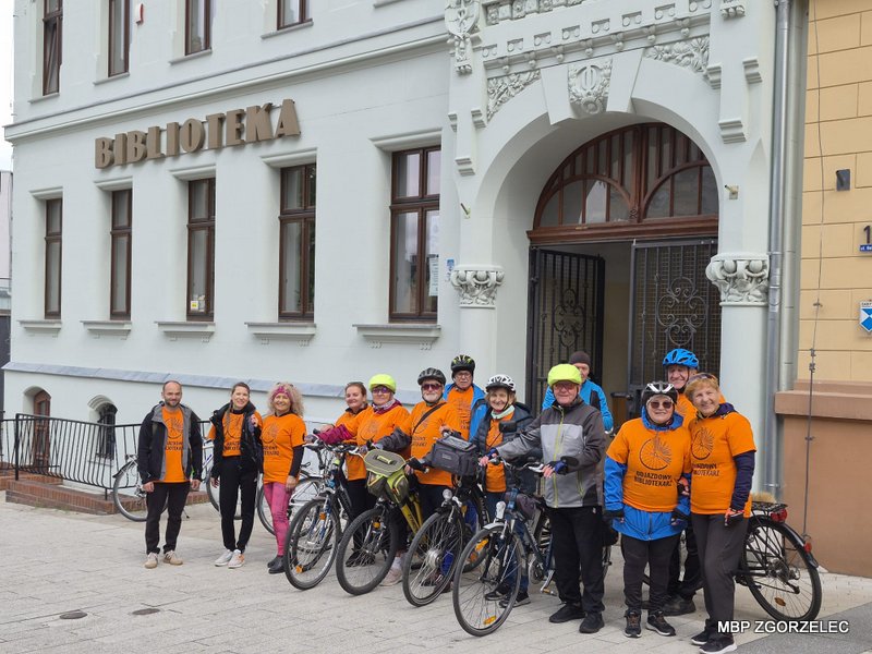 Uczestnicy rajdu rowerowego z rowerami stoją przed budynkiem biblioteki; na elewacji widoczny napis „Biblioteka”.