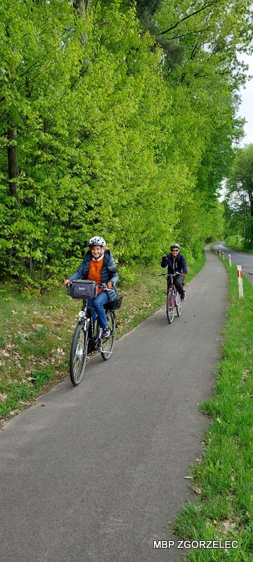 Dwóch uczestników rajdu rowerowego jedzie ścieżką rowerową wśród zieleni; po lewej stronie gęste drzewa, po prawej pobocze i&nbsp;droga lokalna.