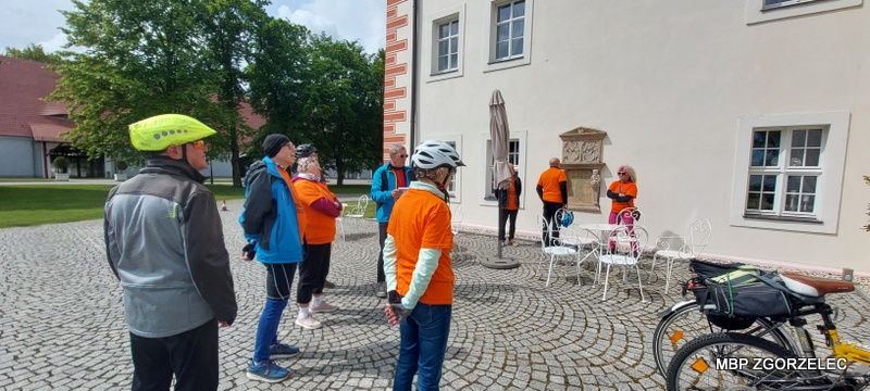Grupa uczestników rajdu rowerowego zatrzymała się na dziedzińcu pałacu; rowery stoją obok, a&nbsp;uczestnicy słuchają informacji o&nbsp;zwiedzanym miejscu.