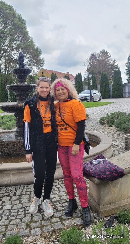 Uczestnicy rajdu rowerowego w&nbsp;pomarańczowych koszulkach stoją przy kamiennej fontannie na dziedzińcu zabytkowego obiektu; w&nbsp;tle widoczna zieleń i&nbsp;zabudowania.