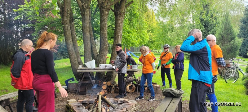 Przerwa podczas rajdu rowerowego – uczestnicy zgromadzeni przy ognisku i&nbsp;stołach piknikowych na terenie zielonym; w&nbsp;tle widoczne drzewa i&nbsp;zaparkowane rowery.
