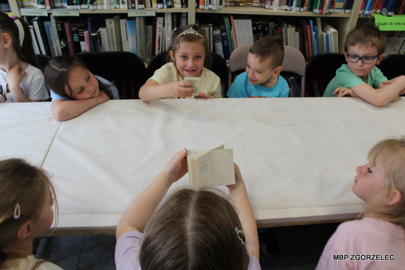 Przedszkolaki siedzą przy stole w&nbsp;bibliotece i&nbsp;oglądają niewielką książkę trzymaną przez jedno z&nbsp;dzieci. W&nbsp;tle znajdują się regały z&nbsp;księgozbiorem