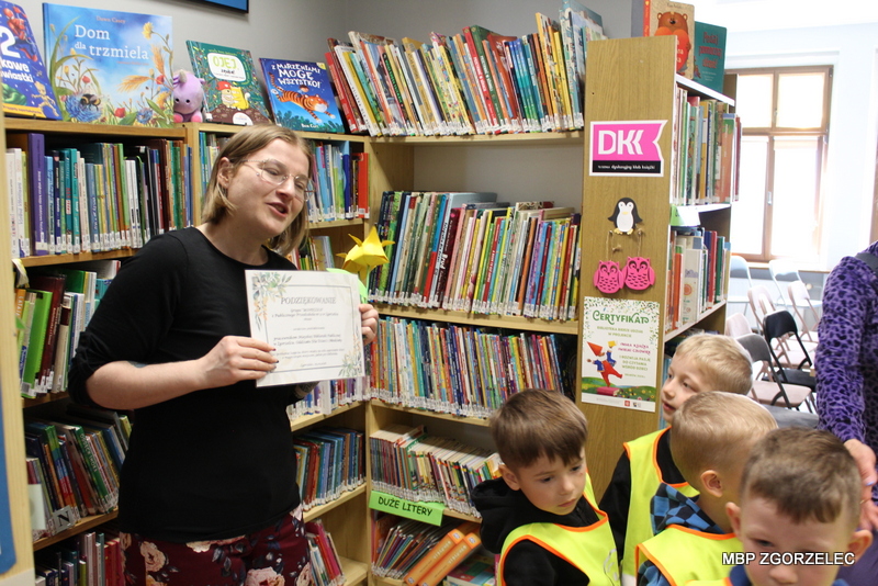 Bibliotekarka stoi w Oddziale dla Dzieci itrzyma podziękowanie, które otrzymała od dzieci podczas spotkania z przedszkolakami. W tle widoczne są regały z książkami dziecięcymi oraz oznaczenia kampanii czytelniczej. Bibliotekarka stoi w Oddziale dla Dzieci itrzyma podziękowanie, które otrzymała od dzieci podczas spotkania z przedszkolakami. W tle widoczne są regały z książkami dziecięcymi oraz oznaczenia kampanii czytelniczej.