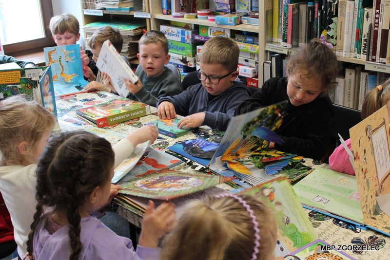 Dzieci siedzą przy stole w&nbsp;bibliotece i&nbsp;wspólnie oglądają książki dla najmłodszych, w&nbsp;tym książki trójwymiarowe pop‑up. W&nbsp;tle regały z&nbsp;księgozbiorem.