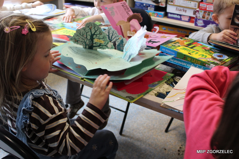 Przedszkolaki siedzą przy stole w bibliotece i oglądają rozłożoną trójwymiarową książkę pop‑up z ilustracjami. Na stole leżą także inne książki dla dzieci, w tle widoczne są gry planszowe na półkach. Przedszkolaki siedzą przy stole w bibliotece i oglądają rozłożoną trójwymiarową książkę pop‑up z ilustracjami. Na stole leżą także inne książki dla dzieci, w tle widoczne są gry planszowe na półkach.