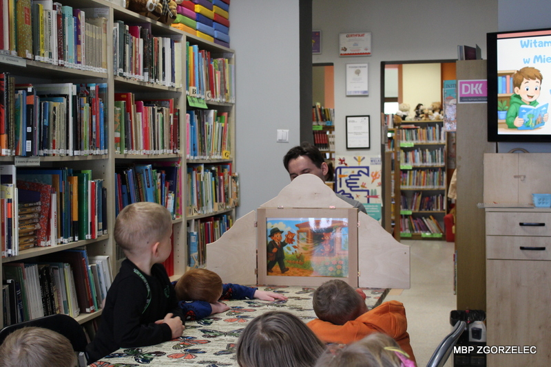 Przedszkolaki siedzą przy stole w bibliotece i oglądają teatrzyk kamishibai prezentowany z drewnianej skrzynki. W tle znajdują się regały z książkami oraz przestrzeń Oddziału dla Dzieci. Przedszkolaki siedzą przy stole w bibliotece i oglądają teatrzyk kamishibai prezentowany z drewnianej skrzynki. W tle znajdują się regały z książkami oraz przestrzeń Oddziału dla Dzieci.