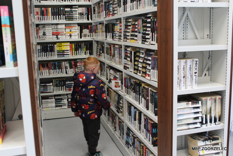 Dziecko stoi między regałami w bibliotece i ogląda książki ustawione na półkach. Widoczny jest korytarz między regałami z księgozbiorem. Dziecko stoi między regałami w bibliotece i ogląda książki ustawione na półkach. Widoczny jest korytarz między regałami z księgozbiorem.