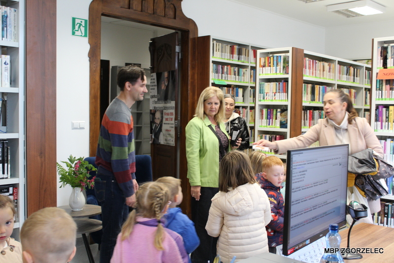 Grupa przedszkolaków wraz z opiekunami stoi w Wypożyczalni dla Dorosłych i słucha informacji podczas zwiedzania biblioteki. W tle widoczne są wysokie regały z książkami. Grupa przedszkolaków wraz z opiekunami stoi w Wypożyczalni dla Dorosłych i słucha informacji podczas zwiedzania biblioteki. W tle widoczne są wysokie regały z książkami.