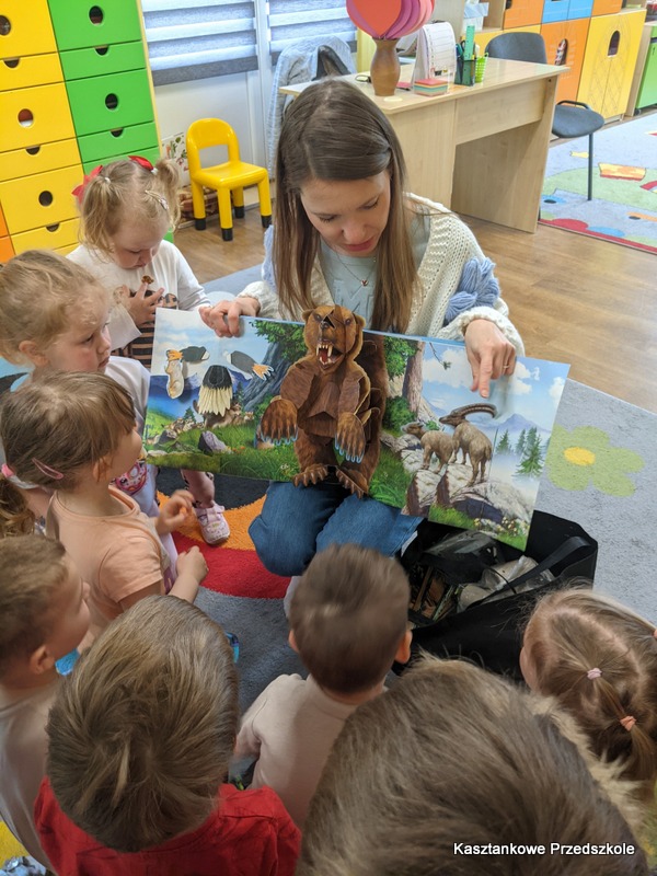 Bibliotekarka siedzi na dywanie w&nbsp;sali przedszkolnej i&nbsp;prezentuje dzieciom trójwymiarową książkę pop‑up z&nbsp;ilustracjami zwierząt. Dzieci siedzą wokół i&nbsp;oglądają książkę.