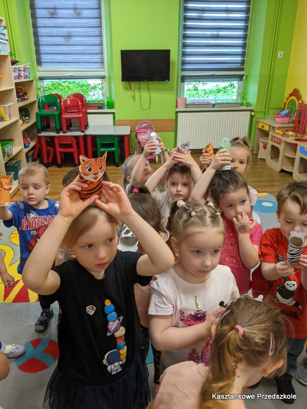 Grupa przedszkolaków stoi w&nbsp;sali przedszkolnej i&nbsp;trzyma w&nbsp;górze kartonowe ilustracje tygrysów. W&nbsp;tle widoczne są półki z&nbsp;zabawkami i&nbsp;meblami.