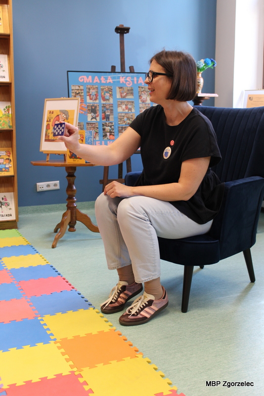 Agnieszka Błazy, prowadząca warsztaty czytelnicze, pokazuje dzieciom kartę do zbierania naklejek podczas wizyt w&nbsp;bibliotece. Na podłodze znajduje się kolorowa mata edukacyjna, a&nbsp;w tle przygotowana przestrzeń biblioteczna dla najmłodszych.
