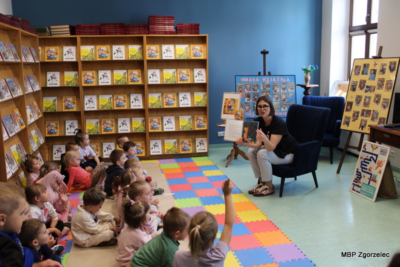 Agnieszka Błazy, prowadząca warsztaty czytelnicze, pokazuje dzieciom ilustrację z&nbsp;książki „Bobuś”. Na podłodze znajduje się kolorowa mata edukacyjna, a&nbsp;w tle przygotowana przestrzeń biblioteczna dla najmłodszych.