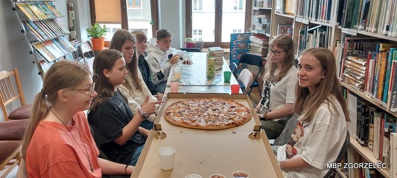 Grupa uczestników spotkania Młodzieżowego Klubu Książki siedzi przy długim stole w&nbsp;bibliotece. Na stole znajduje się duża pizza oraz kubki z&nbsp;napojami. Wokół widoczne są półki z&nbsp;książkami, charakterystyczne dla przestrzeni bibliotecznej.