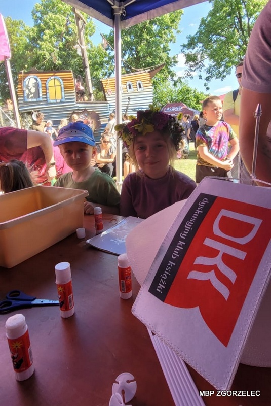 Dwoje dzieci siedzi przy stole warsztatowym z&nbsp;materiałami plastycznymi; widoczny znak DKK oraz stanowisko zajęć w&nbsp;plenerze.
