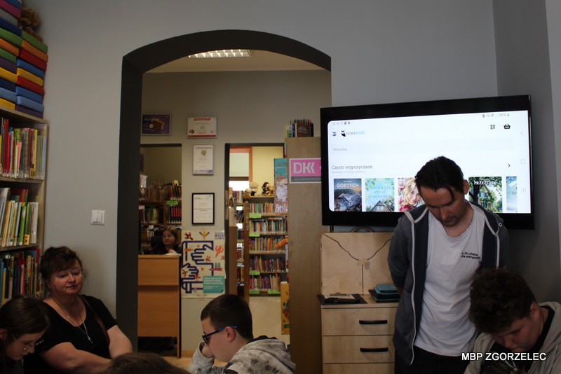 Wnętrze oddziału dziecięco-młodzieżowego biblioteki – grupa uczniów siedzi i&nbsp;stoi wokół prowadzącego zajęcia. Na ekranie telewizora widoczny jest interfejs katalogu bibliotecznego SowaMobi z&nbsp;okładkami książek. W&nbsp;tle regały z&nbsp;książkami i&nbsp;przejście do kolejnego pomieszczenia.