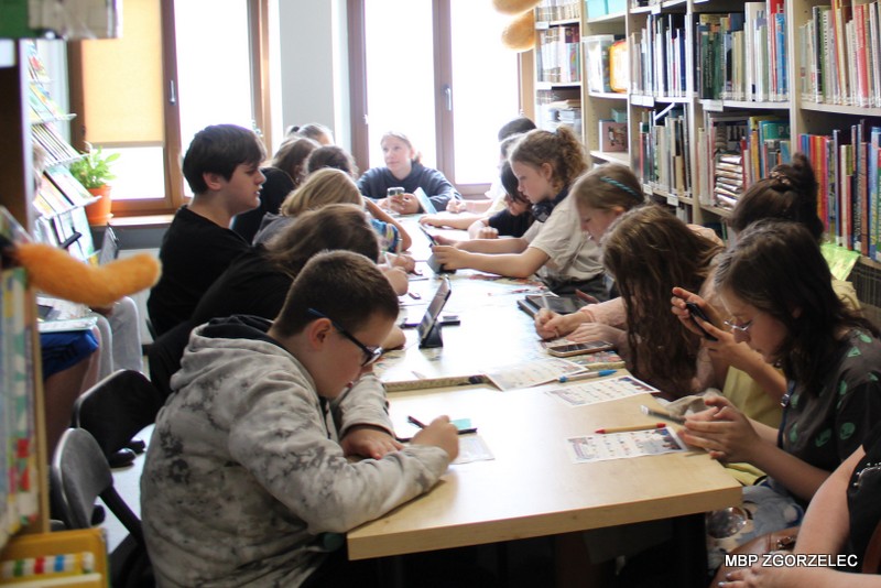 Grupa uczniów siedzi przy długim stole w&nbsp;bibliotece i&nbsp;pracuje nad zadaniami. Uczniowie korzystają z&nbsp;telefonów i&nbsp;kart pracy. W&nbsp;tle widoczne są wysokie regały z&nbsp;książkami oraz duże okno doświetlające pomieszczenie.