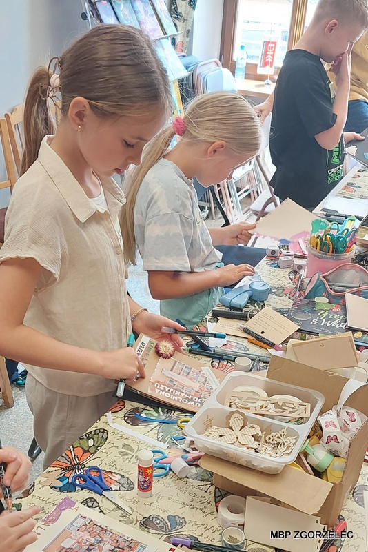 Uczestnicy warsztatów scrapbookingowych stoją przy stole w&nbsp;bibliotece i&nbsp;wycinają oraz dekorują elementy do swoich albumów „Księga moich marzeń”; na stole widoczne są nożyczki, kleje, taśmy ozdobne oraz wycinki z&nbsp;czasopism i&nbsp;książek przygotowane do pracy twórczej.