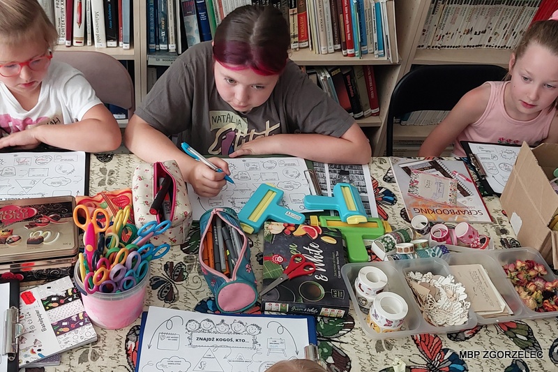 Uczestnicy warsztatów scrapbookingowych siedzą przy stole w&nbsp;bibliotece; na stole widoczne są nożyczki, taśmy ozdobne, stemple oraz wycinki z&nbsp;czasopism i&nbsp;książek.