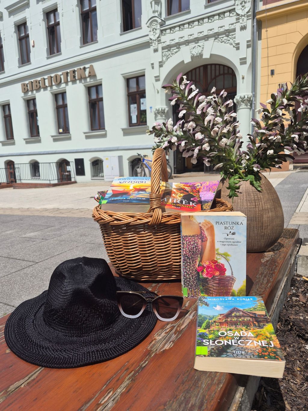 Na drewnianej ławce przed budynkiem biblioteki ustawiono letnią aranżację z książkami. W wiklinowym koszyku znajdują się powieści obyczajowe, a obok stoją dwie książki wyeksponowane okładkami do przodu. Przed koszykiem leżą czarny kapelusz i okulary przeciwsłoneczne. Z prawej strony ustawiono duży wazon z kwitnącymi roślinami. W tle widoczna jest jasna elewacja budynku z dużym napisem „BIBLIOTEKA” oraz ozdobnym wejściem. Zdjęcie utrzymane jest w letnim, słonecznym klimacie i zachęca do wakacyjnej lektury. Zdjęcie jest odnośnikiem do wpisu o otwartej sobocie w Wypożyczalni.