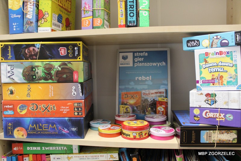 Na regale w Miejskiej Bibliotece Publicznej w Zgorzelcu prezentowana jest kolekcja gier planszowych udostępnionych w ramach programu „Zgrane Biblioteki”. Widoczne są pudełka gier, m.in. „Splendor”, „Avel”, „Dixit”, „MLEM: Agencja Kosmiczna”, „Trio”, „Wydry” i „Cortex”. Pośrodku znajduje się tabliczka z napisem „Strefa gier planszowych”, a obok ustawiono puszki z grą „Dobble”. Kolekcja gier jest wyeksponowana na bibliotecznym regale wśród książek. Na regale w Miejskiej Bibliotece Publicznej w Zgorzelcu prezentowana jest kolekcja gier planszowych udostępnionych w ramach programu „Zgrane Biblioteki”. Widoczne są pudełka gier, m.in. „Splendor”, „Avel”, „Dixit”, „MLEM: Agencja Kosmiczna”, „Trio”, „Wydry” i „Cortex”. Pośrodku znajduje się tabliczka z napisem „Strefa gier planszowych”, a obok ustawiono puszki z grą „Dobble”. Kolekcja gier jest wyeksponowana na bibliotecznym regale wśród książek.