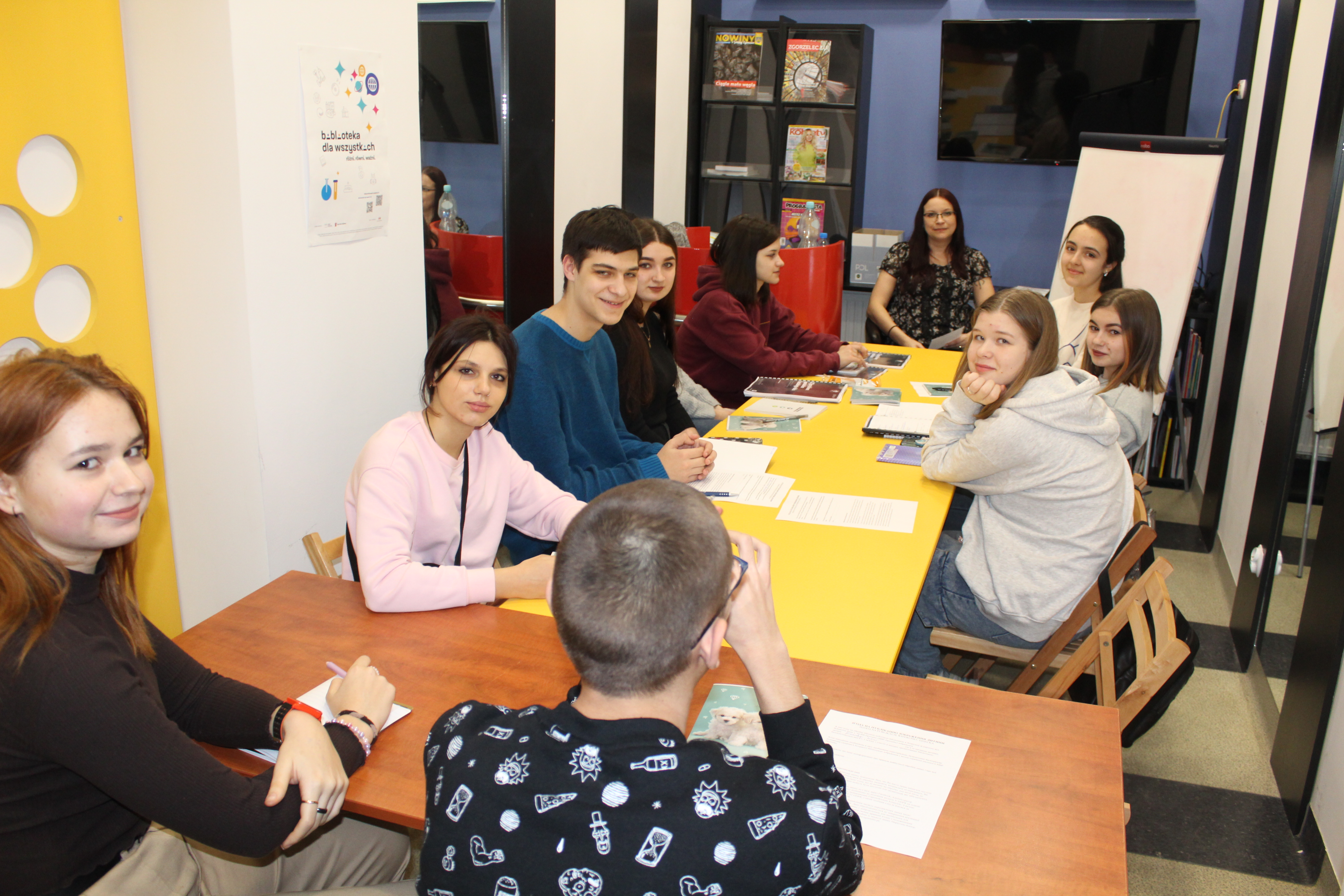 Grupa dziesięciu osób w wieku nastoletnim siedzi przy żółtych stołach w kolorowo urządzonym pomieszczeniu. Na stołach leżą podręczniki do nauki.